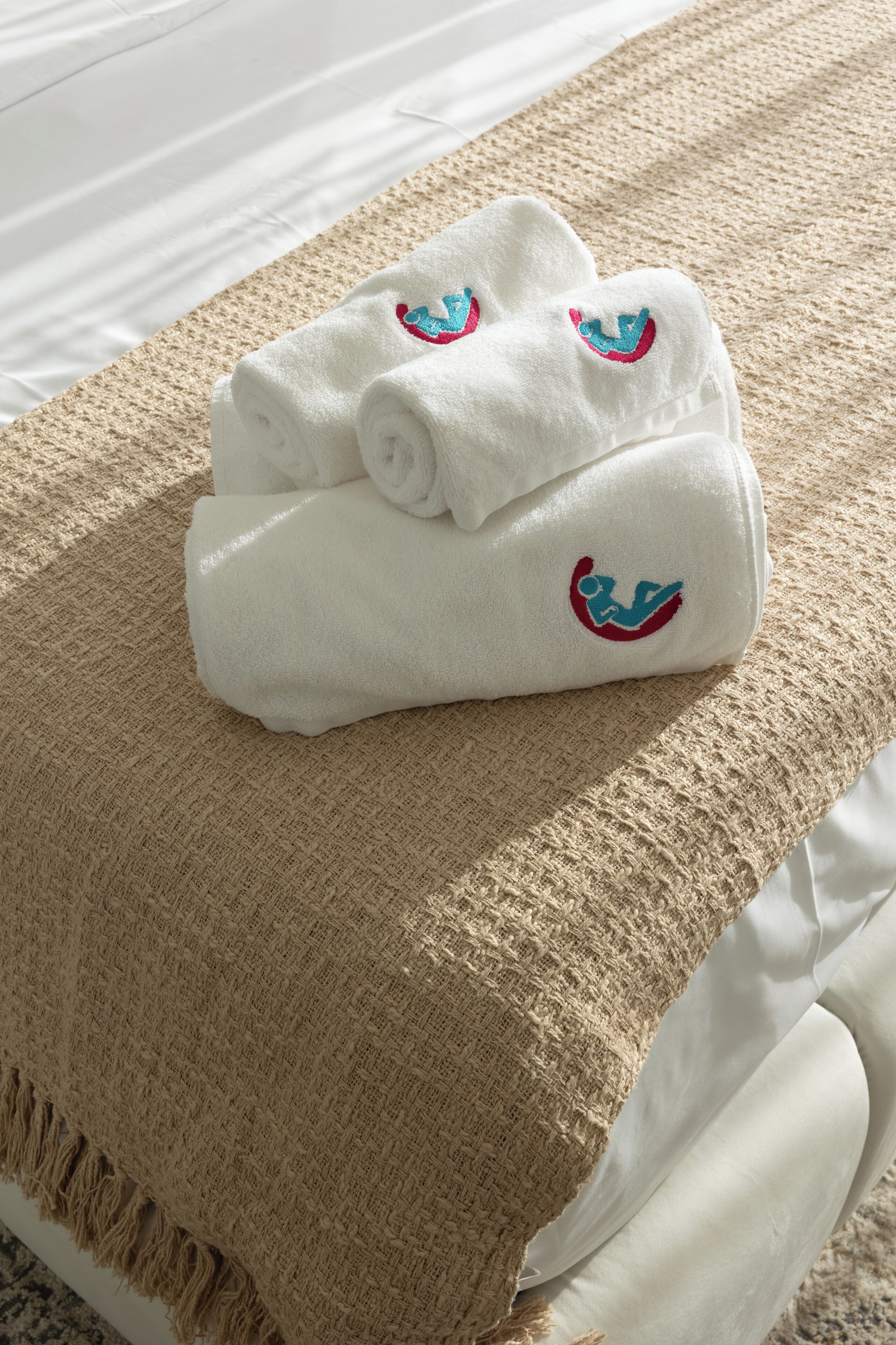 Een keurig gevouwen witte handdoek met een blauw en rood logo, rustend op een beige deken op een bed.