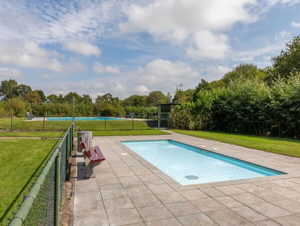 Piscina retangular com um deck ladrilhado, cercada por uma cerca verde e uma área gramada.