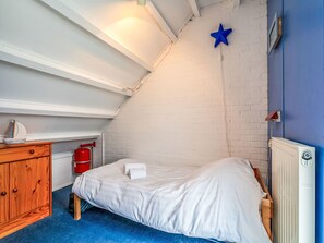 Um quarto pequeno e acolhedor com teto inclinado, uma cama, uma cômoda, um extintor de incêndio vermelho e uma estrela azul decorativa na parede.