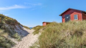 Un sendero arenoso que lleva a una construcción de madera roja con una puerta y una ventana, rodeada de hierba y dunas.