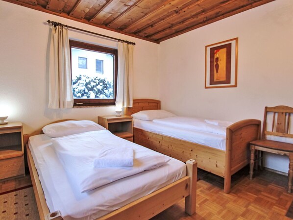 Una habitación con dos camas de madera, una ventana con cortinas, un cuadro en la pared y una silla de madera.