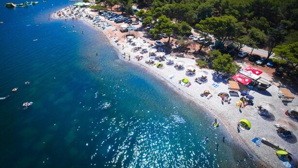 Une vue aérienne d’une plage avec une eau d’un bleu limpide, un rivage sablonneux et de nombreux parasols et tentes de plage.