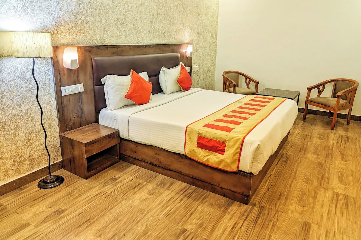 Ein ordentlich eingerichtetes Schlafzimmer mit einem Holzkopfteil, einem Bett mit weißer Bettwäsche und roten Akzenten, zwei Holzstühlen und einem Nachttisch.