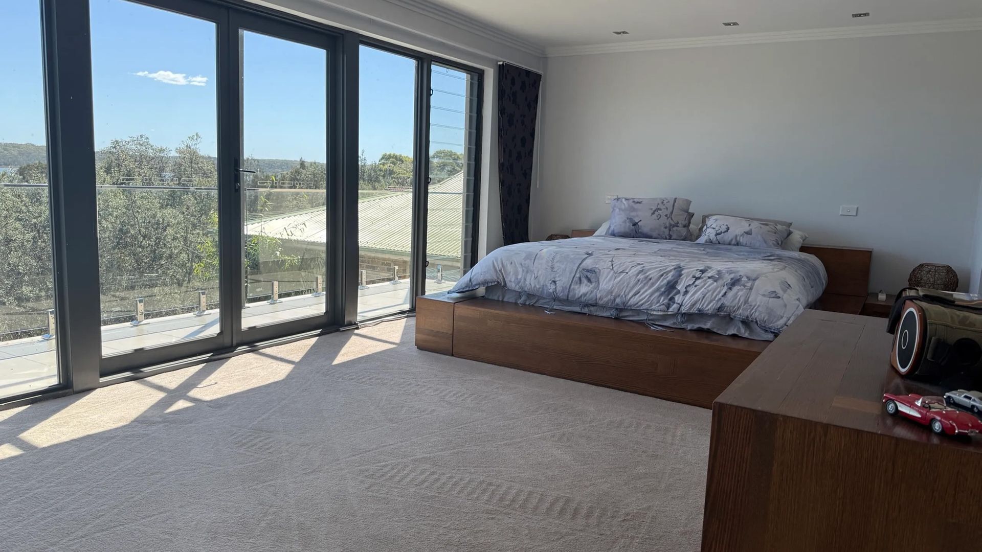 Une chambre avec une grande fenêtre, un lit recouvert d’une couette à motifs floraux et une table de chevet en bois.