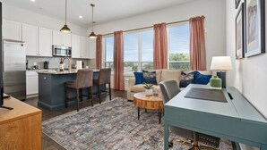 Un appartement moderne comprenant une cuisine, un coin repas et un salon. Il est doté d’un grand îlot central dans la cuisine, d’une table à manger avec des chaises, d’un canapé avec des coussins et d’un bureau avec une lampe.