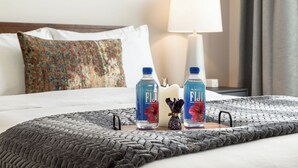 Una cama bien hecha con una manta gris de punto, dos botellas de Fiji, una vela blanca y un pequeño florero decorativo sobre una mesita de noche.