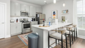 Una cocina moderna con armarios blancos, electrodomésticos de acero inoxidable, una isla central con taburetes y una ventana con persianas.
