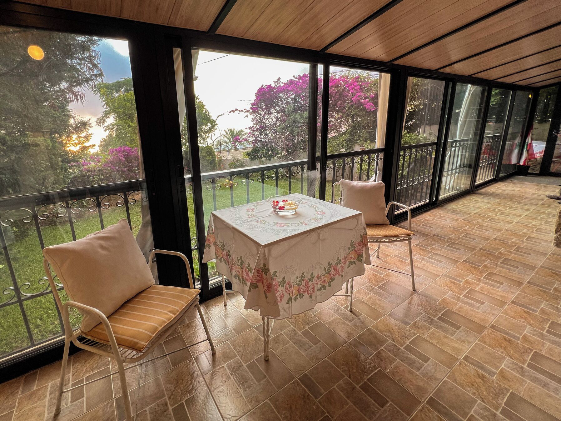 A covered outdoor seating area with a table and chairs, large windows, and a view of the garden.