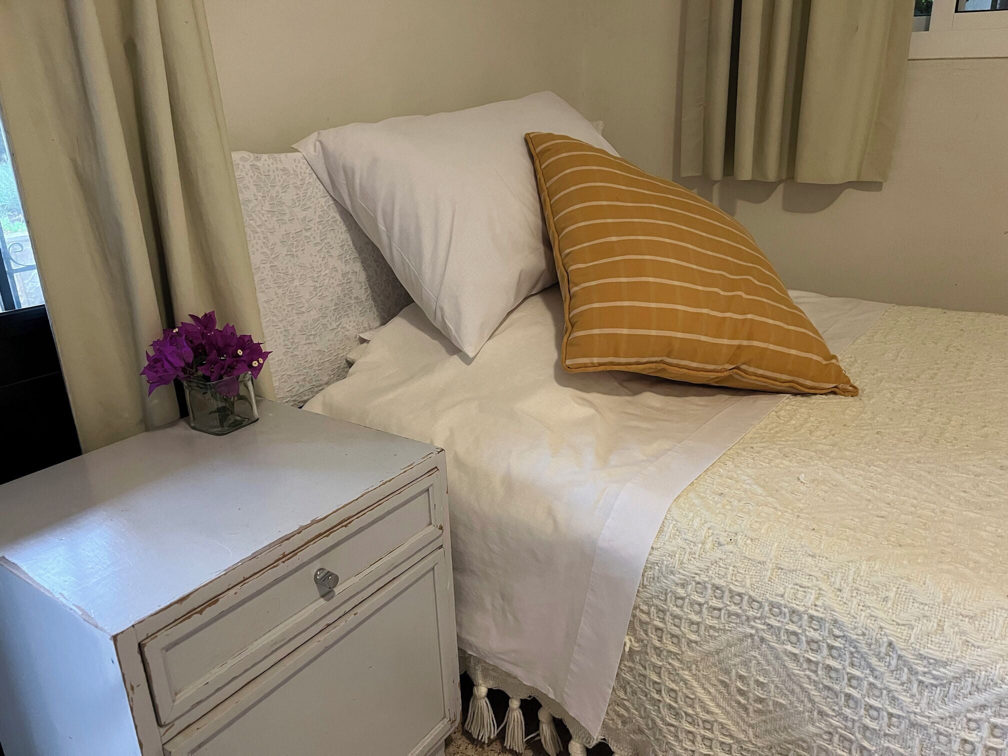 Una cama con ropa de cama blanca y una almohada a rayas, una mesita de noche con un jarrón de flores moradas, y un mesita de noche blanca con un cajón.