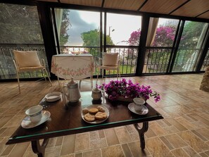 A dining area with a table set for tea, surrounded by chairs, with a view of a garden through large windows. - Les Bougainvilliers GuestHouse by Hansa  (Chemlane)