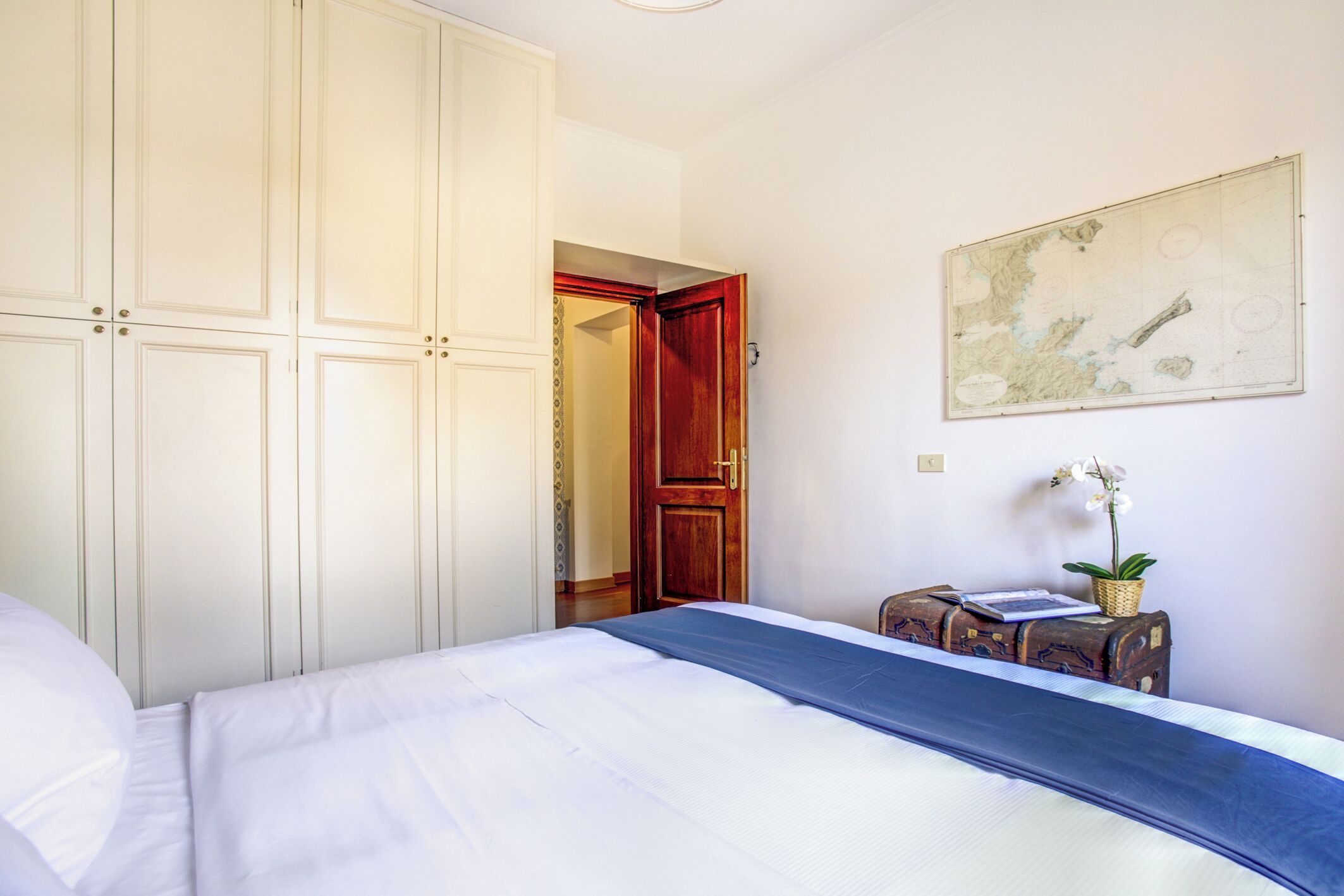 A bedroom with a large bed, a wardrobe, a wooden door, a map on the wall, and a small table with a plant.