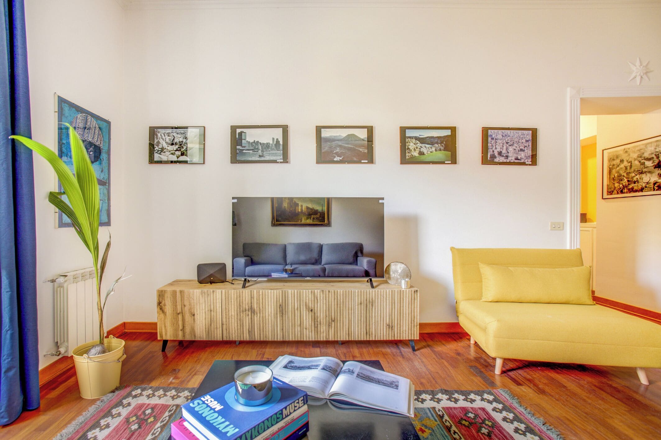 Ein Wohnzimmer mit einer Couch, einem gelben Sessel, einem Couchtisch mit Büchern, einer Pflanze und gerahmten Bildern an der Wand.