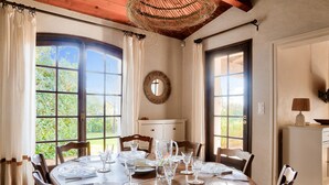 A dining area with a round table set for six, surrounded by wooden chairs. There are large windows with curtains, a wooden ceiling, and a small cabinet with a lamp.
