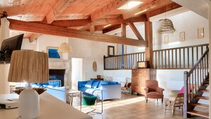 A spacious living area with a high ceiling, wooden beams, a fireplace, a staircase, and a variety of seating options.