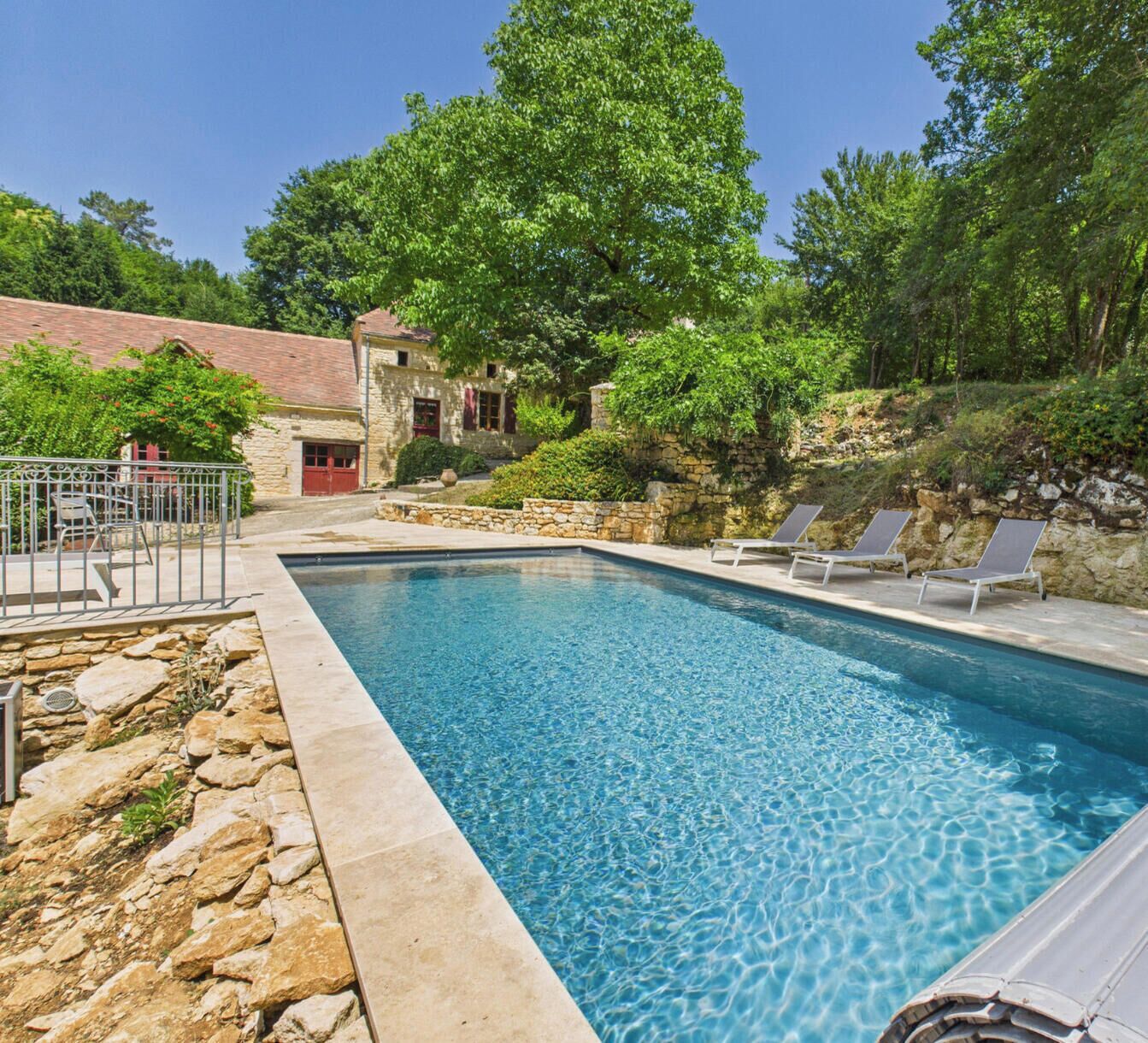 A rectangular swimming pool with clear blue water, surrounded by stone walls and a stone building with a red door.