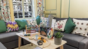 Un salon avec un canapé gris, des coussins colorés, une table basse en bois avec des jouets, une plante en pot et une fenêtre ornée de rideaux à motifs floraux.