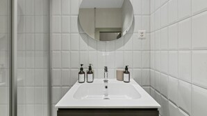 Et moderne badeværelse med hvide fliser, en rektangulær vask, et rundt spejl og tre flasker toiletartikler.