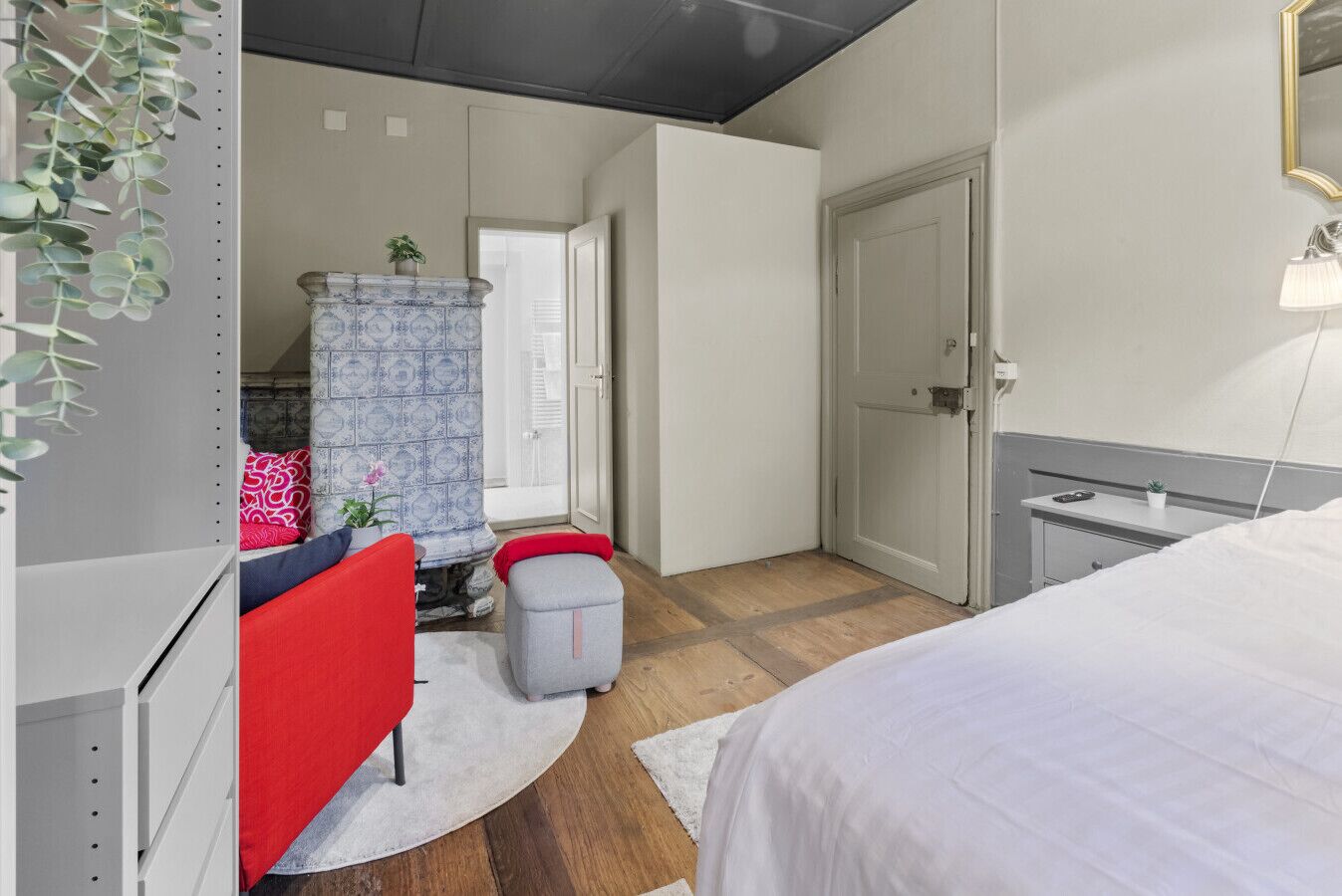 A bedroom with a bed, a red chair, a small table, a grey ottoman, a patterned cabinet, and a window with curtains.