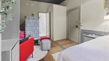 A bedroom with a bed, a red chair, a small table, a grey ottoman, a patterned cabinet, and a window with curtains.