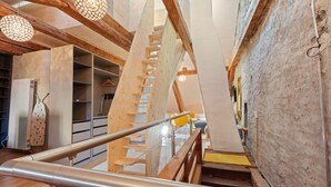 Un escalier en bois avec une rampe métallique, menant à une pièce dont un des murs est en pierre et qui est éclairée par un luminaire suspendu.