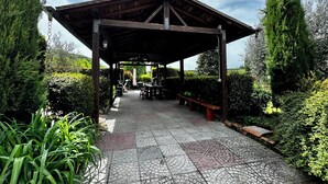 Un'area salotto esterna coperta con tavoli e panche, immersa nel verde e accessibile tramite un viale lastricato.