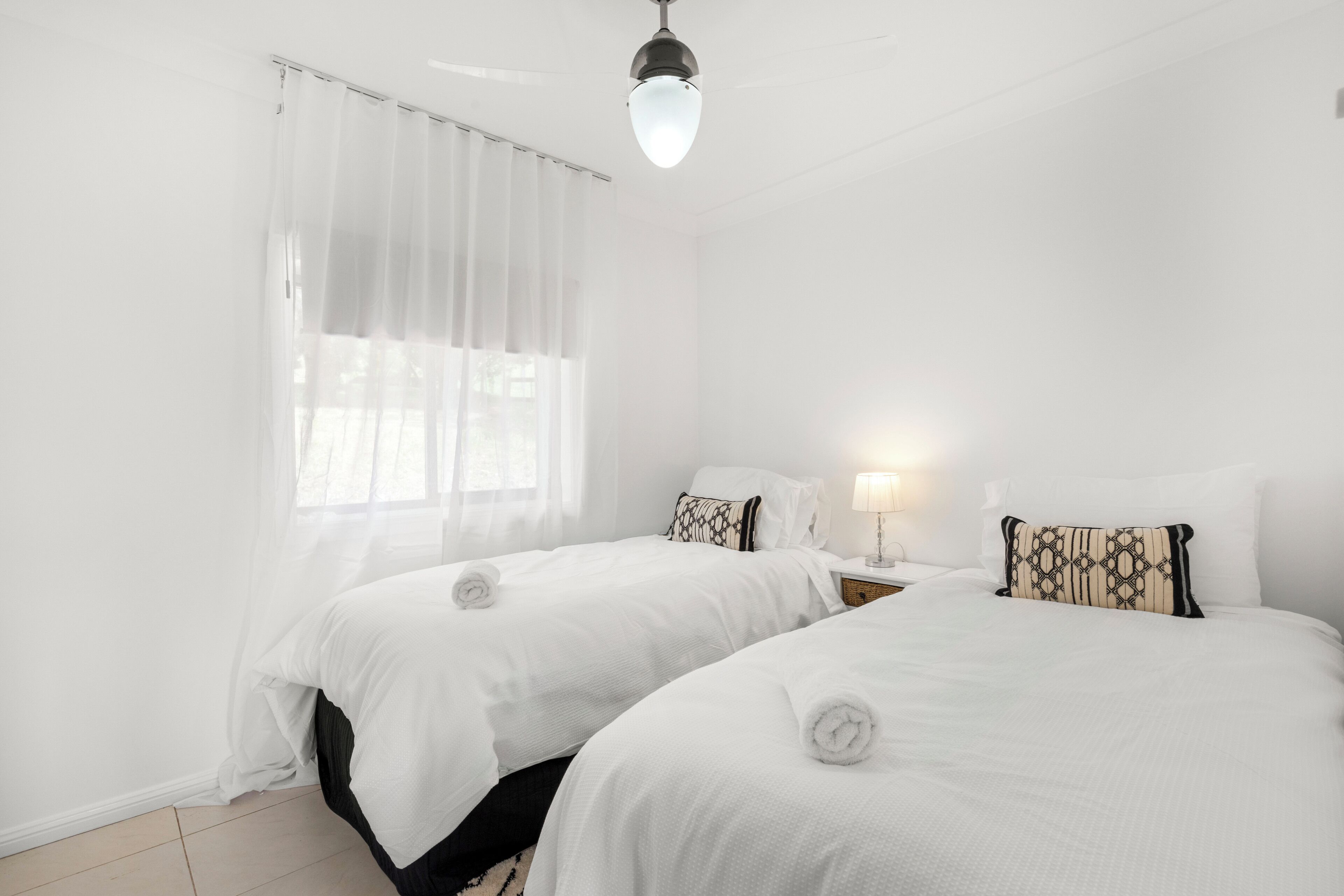 A hotel room with two beds, white bedding, a window with sheer curtains, a bedside table with a lamp, and a ceiling light.
