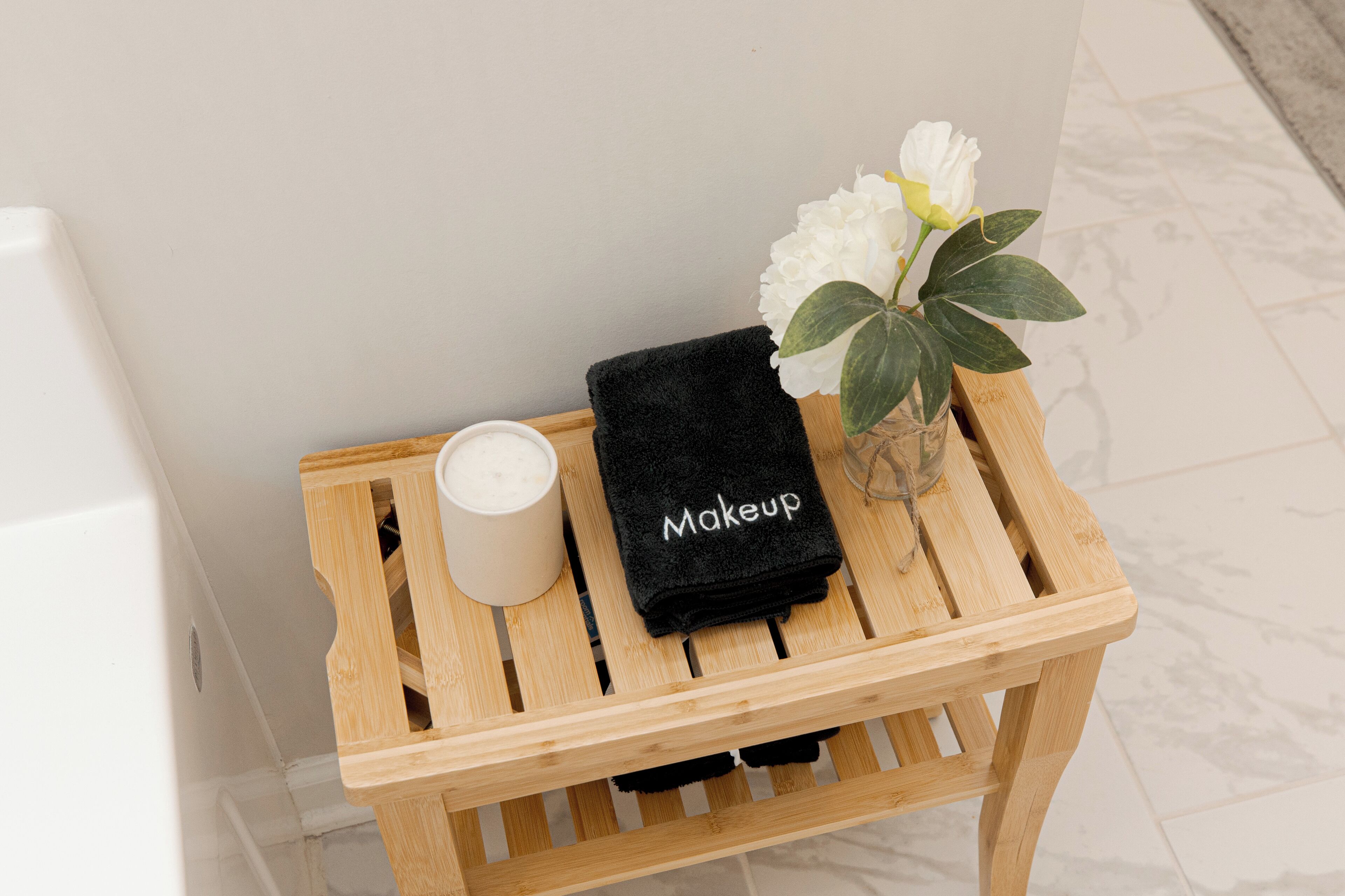 Un taburete de madera con una vela blanca, una toalla negra que dice "Makeup" y un jarrón con flores blancas.