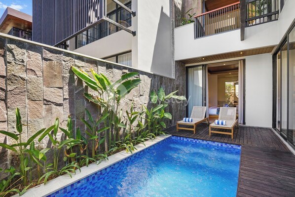 A modern outdoor pool area with a stone wall, tropical plants, and two lounge chairs.