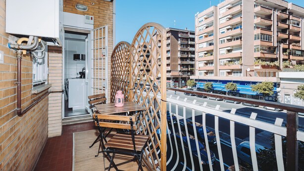 A balcony with a wooden table and chairs, a view of a city street, and a building with multiple balconies.
