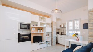 A modern kitchen with white cabinetry, a built-in oven, and a microwave. There is a dining area with a white table and blue sofa.
