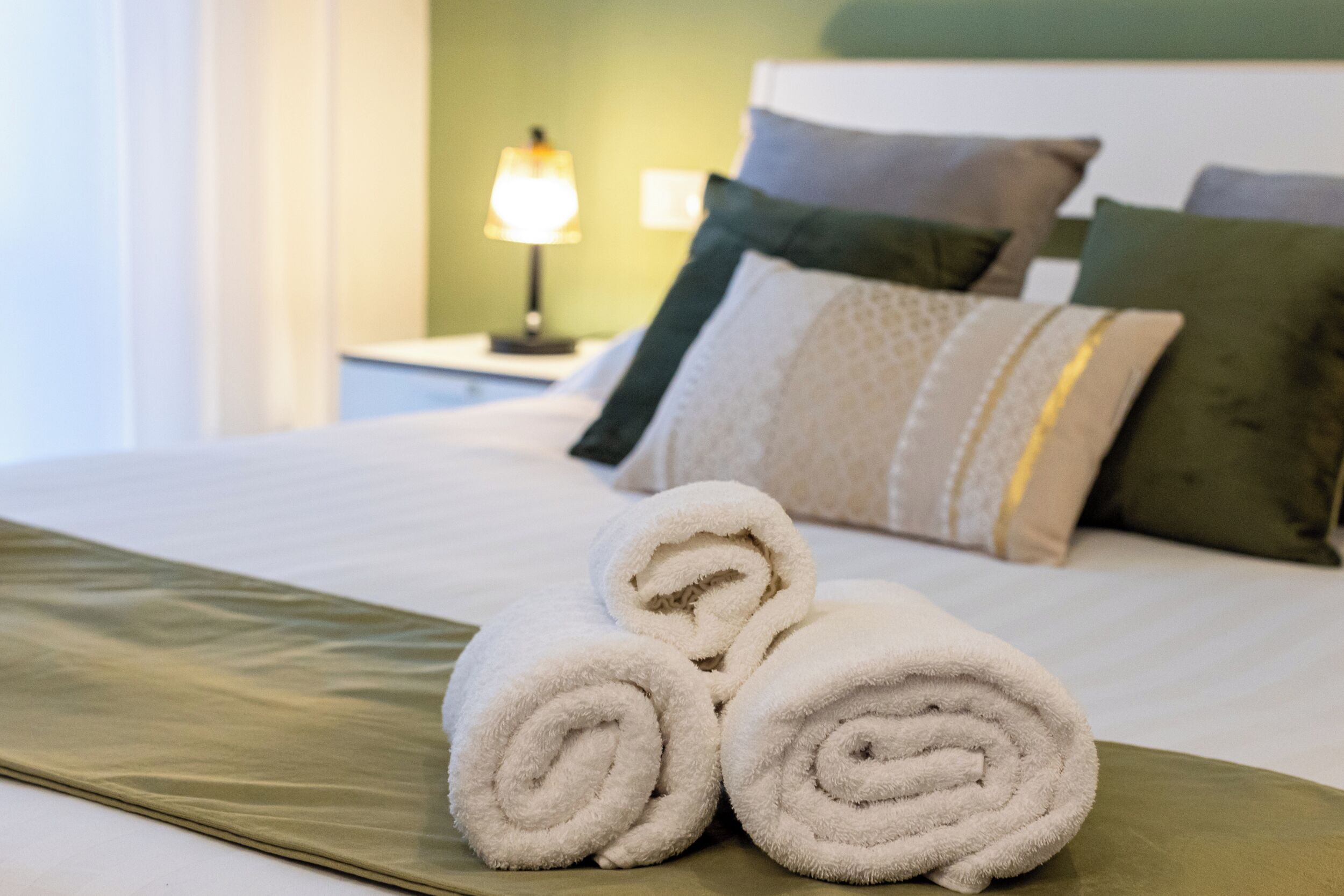 A neatly made bed with rolled white towels, a green pillow, and a lamp on a bedside table.
