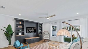 A modern living room with a sofa, chairs, a coffee table, a TV, and a large window with curtains. - Blue Wave by Team Joseph Ellen (Jacksonville Beach)