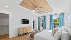 A bedroom with a bed, a ceiling fan, a window with curtains, a wooden cabinet, and a sign on the wall. - Blue Wave by Team Joseph Ellen (Jacksonville Beach)