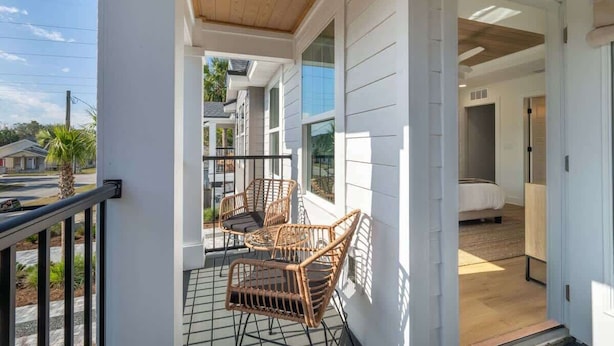 A balcony with wicker furniture, a view of the street, and a bedroom with a bed and nightstand. - Blue Wave by Team Joseph Ellen (Jacksonville Beach)