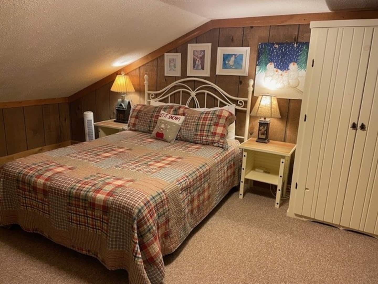 A bedroom with a bed, two bedside tables, a nightstand, a wardrobe, and a sloped ceiling.