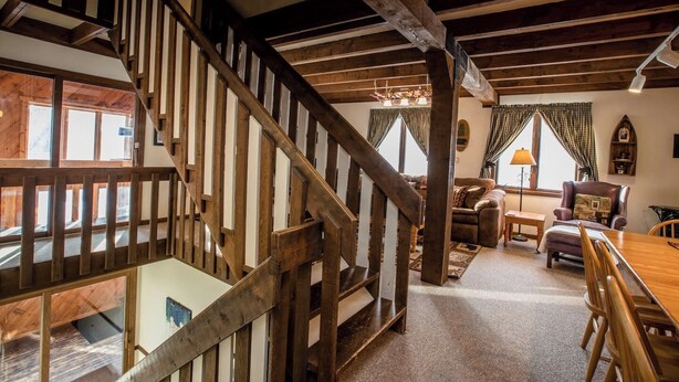 A wooden staircase leading to an upper floor, a cozy living room with a sofa, a table, and a chair, and a view of the outside through large windows. - A Wonderful Vacation Home Set in the Woods with Unique Envelope Architecture (Bessemer)