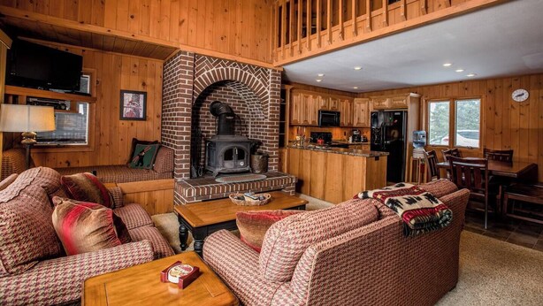 A cozy cabin interior with a fireplace, wooden furniture, a kitchen area, and a dining space.