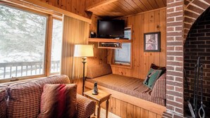 A cozy cabin interior with wooden walls, a fireplace, a television, and a window overlooking a snowy landscape.