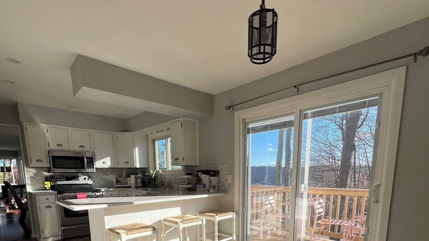 Et moderne køkken med hvide skabe, en mikrobølgeovn og komfur. Der er et spiseområde med bord og stole, samt en balkon med udsigt til træer.