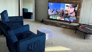 En stue med et stort fladskærms-tv monteret på en murstensvæg, en blå sofa, en lille skammel og en trampolin.