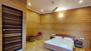Une chambre avec un plancher en bois, un lit recouvert de draps blancs, une table de chevet, une chaise rouge et un ventilateur de plafond.