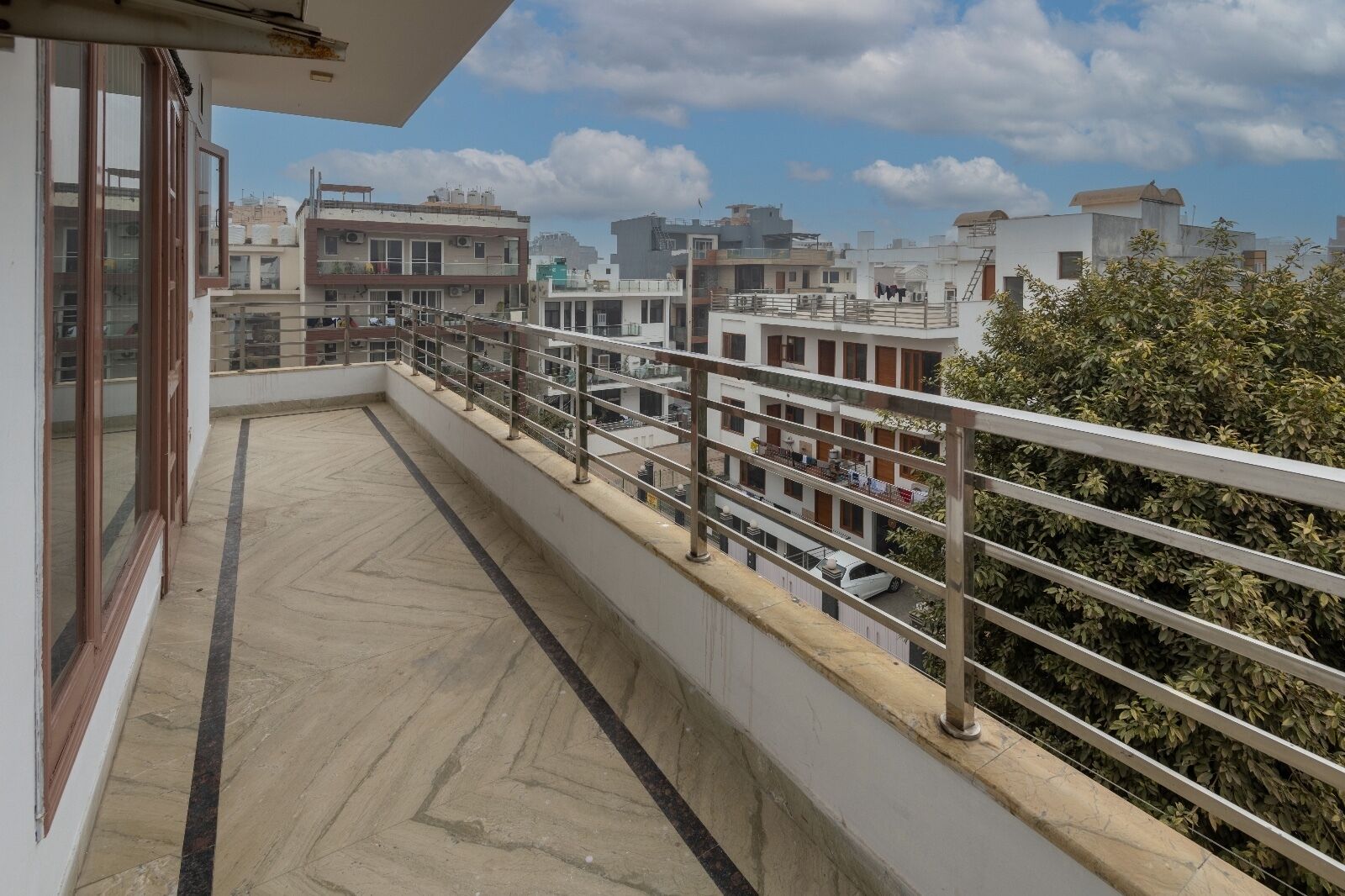 A balcony with a tiled floor and metal railing, overlooking a residential area with multiple buildings and a clear sky.