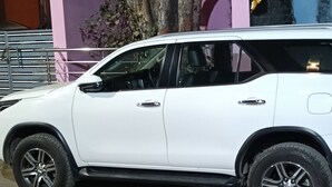 Un SUV blanco estacionado frente a un edificio con un árbol, un muro morado y un pilar rosa.