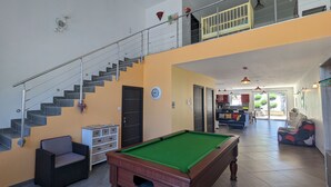 A spacious living area with a pool table, a sofa, a staircase, and a kitchen in the background.