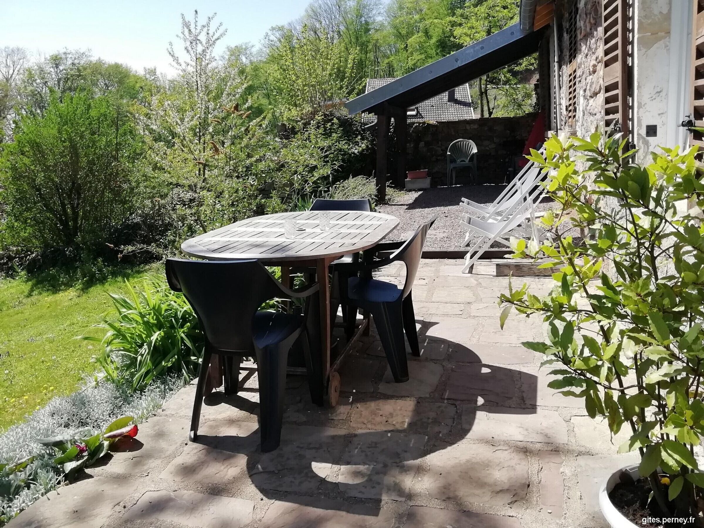 An outdoor patio with a round table and chairs, surrounded by greenery and a stone wall.