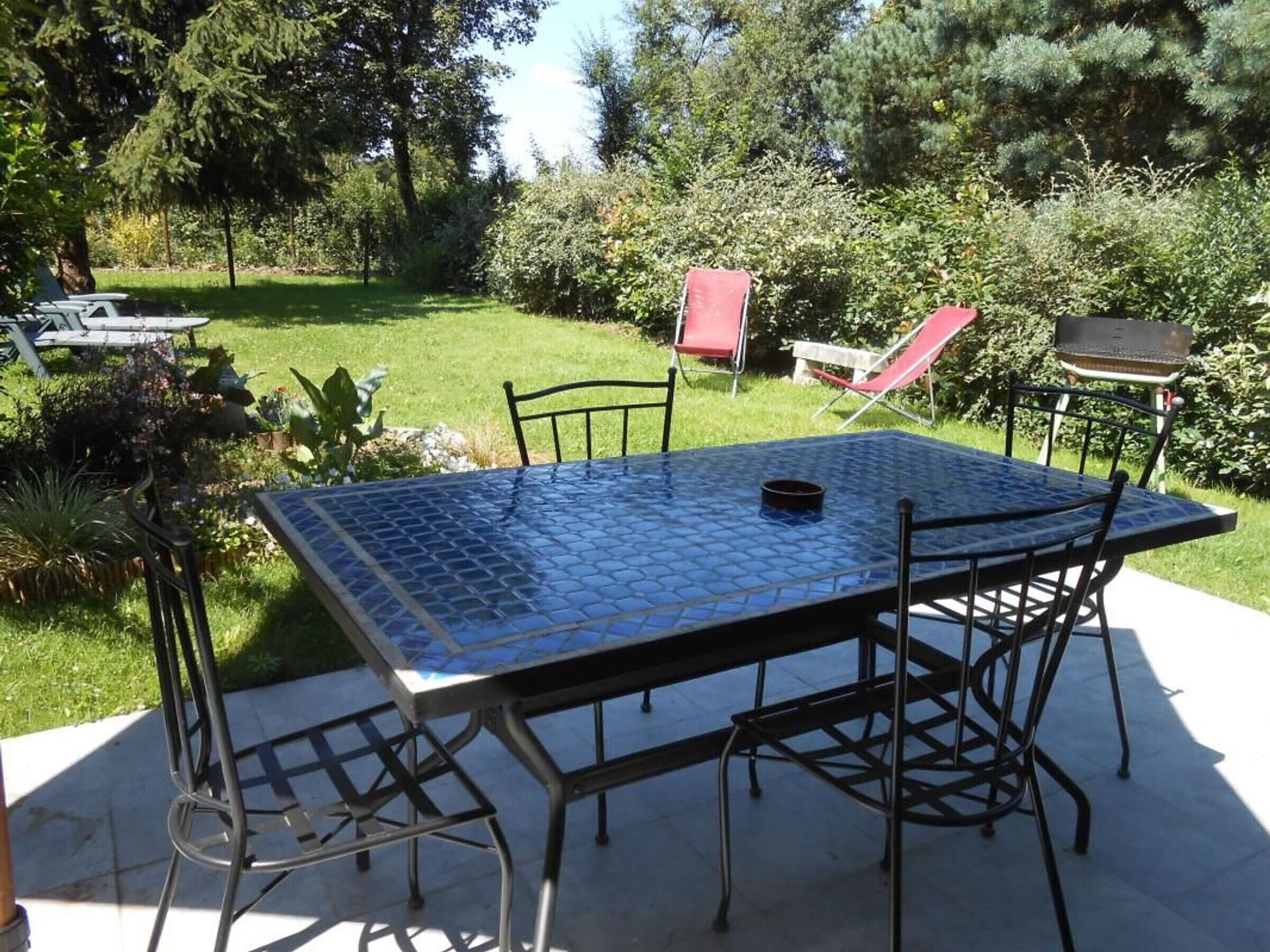 Une terrasse avec une table carrée et des chaises, entourée de verdure et agrémentée d’un barbecue.