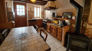 Una cocina con armarios de madera, suelo de baldosas, una mesa de comedor con sillas, una estufa y una chimenea negra.