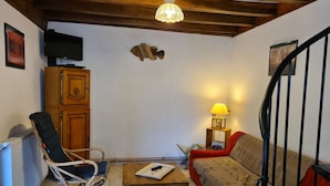 Una sala de estar acogedora con techo de madera, un televisor montado en la pared, un mueble de madera, un sillón de mimbre, un sofá rojo y una pequeña mesa con una lámpara.
