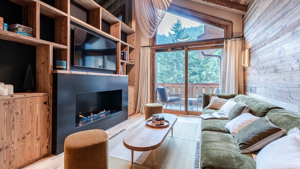 A cozy living room with a fireplace, wooden shelves, and a large window overlooking a deck.