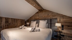 Um quarto com teto inclinado, painéis de madeira, uma cama grande com roupa de cama branca, duas luminárias de cabeceira e uma pequena mesa com abajur.
